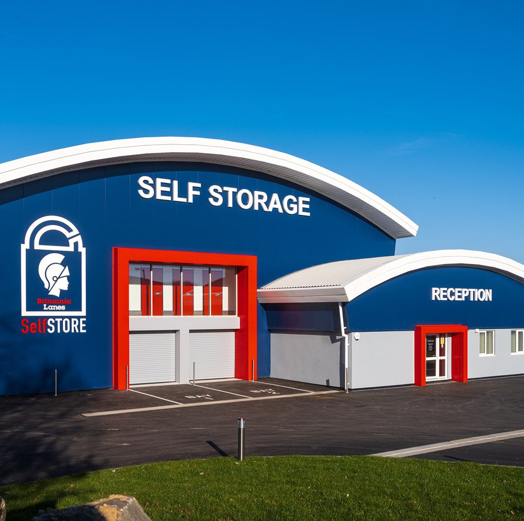 LOC2-323-min A blue and white self-storage facility with large signage reading "SELF STORAGE" and "RECEPTION." The building has red accents around the entrances and a logo featuring a lock and helmeted figure. The area is paved with a clear, blue sky above.